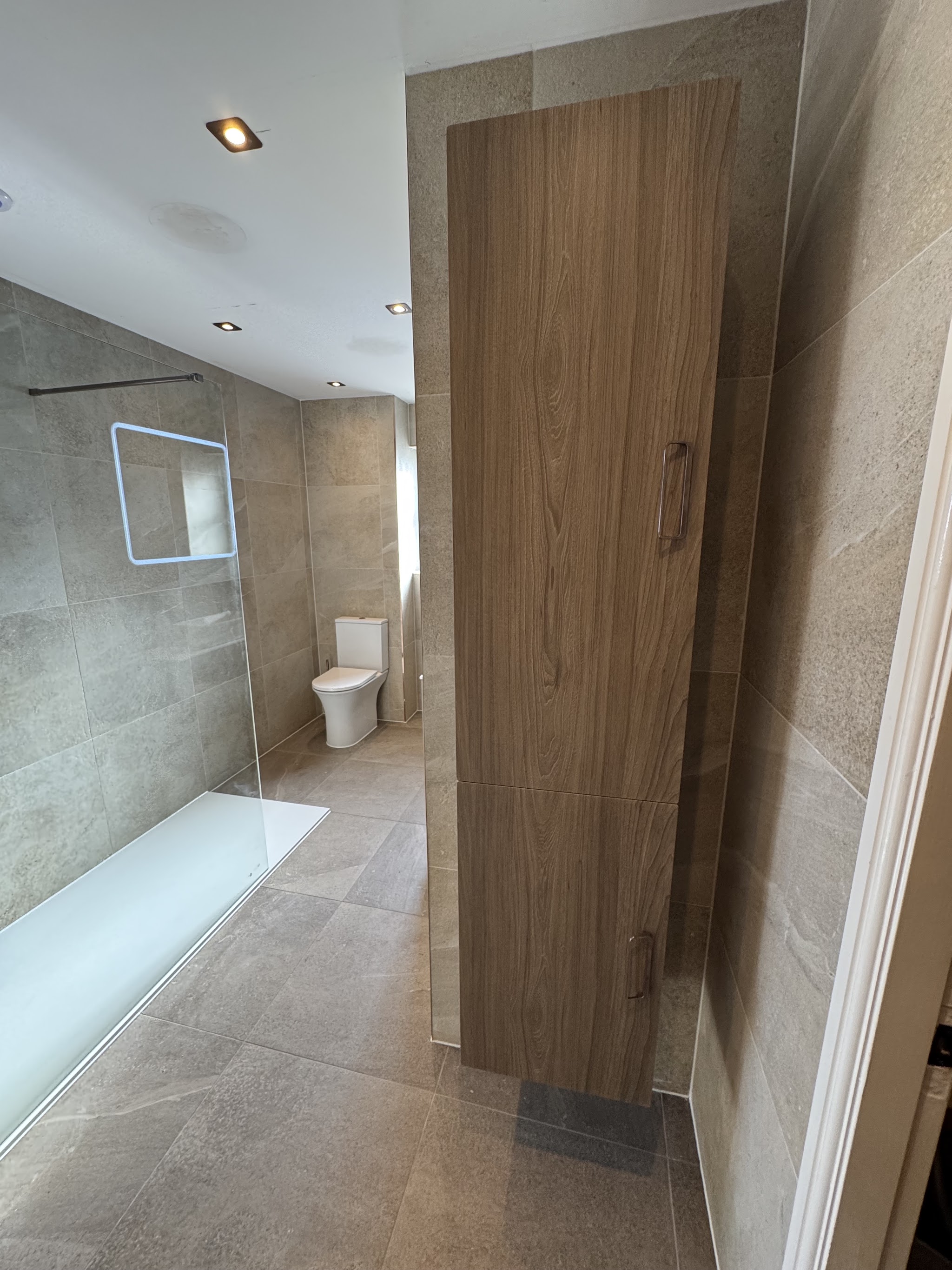 Tall timber storage unit in a tiled bathroom with a walk-in shower and backlit mirror behind